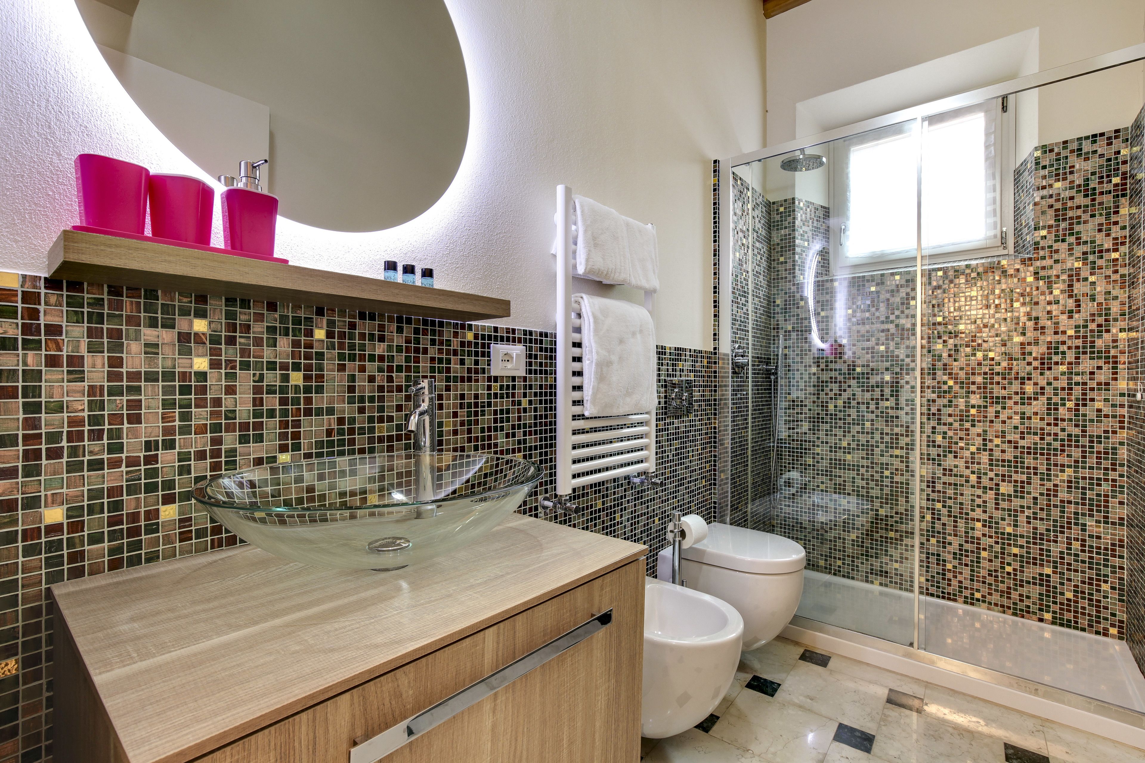 Et moderne badeværelse med vask i glas, toilet og bruseområde med mosaikfliser.