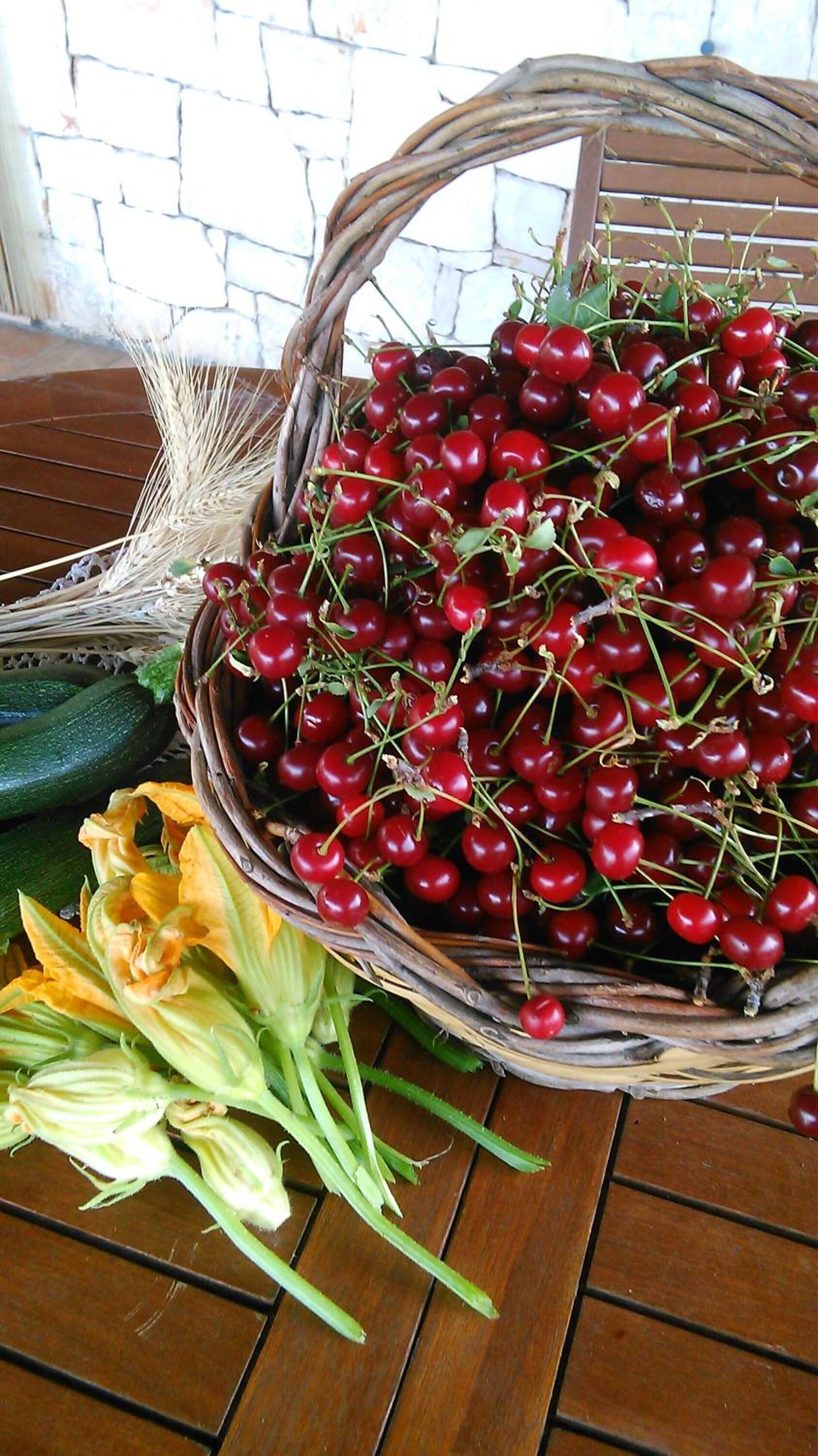 A wicker basket filled with cherries, a cucumber, and some flowers on a wooden table.
