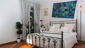 A bedroom with a metal bed frame, white bedding, a colorful quilt, a potted plant, and a floor lamp. - Stupor Mundi (Altamura)