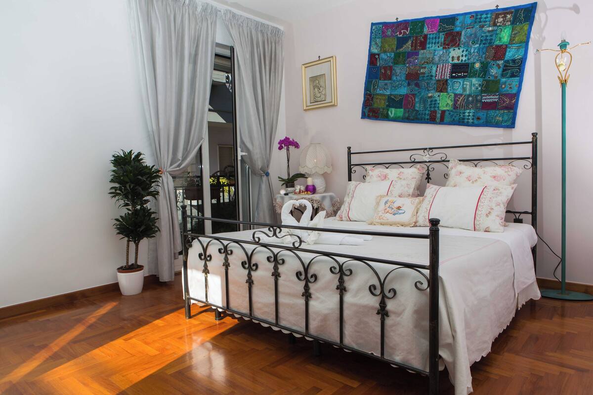 A bedroom with a metal bed frame, white bedding, a colorful quilt, a potted plant, and a floor lamp.