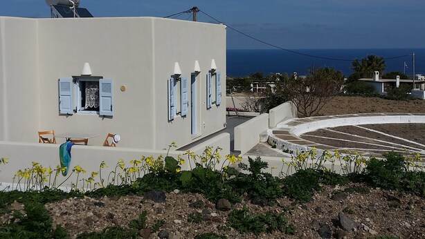 A white building with blue shutters, a small garden with yellow flowers, and a view of the sea. - Theros Villa (Santorini)