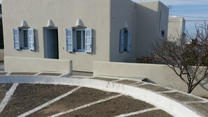 A building with white walls and blue shutters, a paved area with white lines, and a tree with bare branches. - Theros Villa (Santorini)