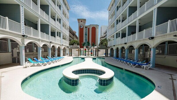 A pool area with lounge chairs, a central water feature, and a building with balconies. - Myrtle Beach Villas 302A - 6 Bedroom Condo (Myrtle Beach)