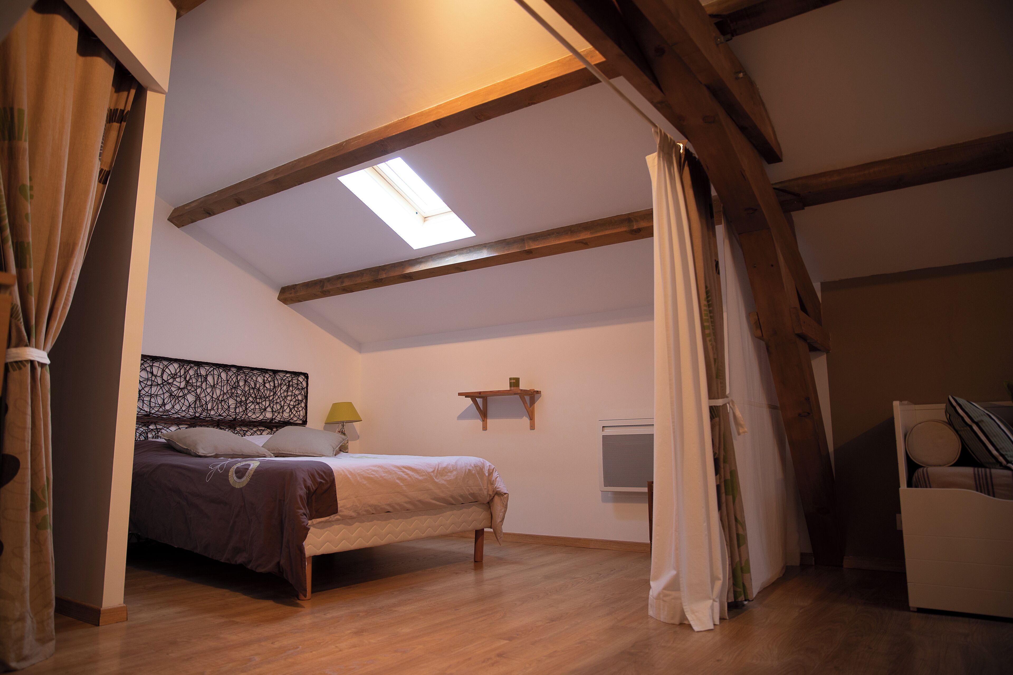 Een slaapkamer met een bed, houten balken, een dakraam, een nachtkastje en een klein plankje.