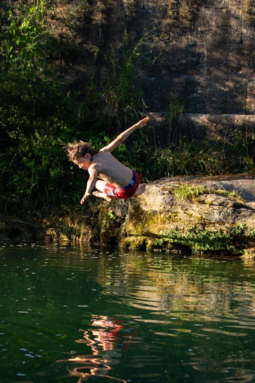 Une personne qui saute dans une étendue d’eau entourée de verdure.