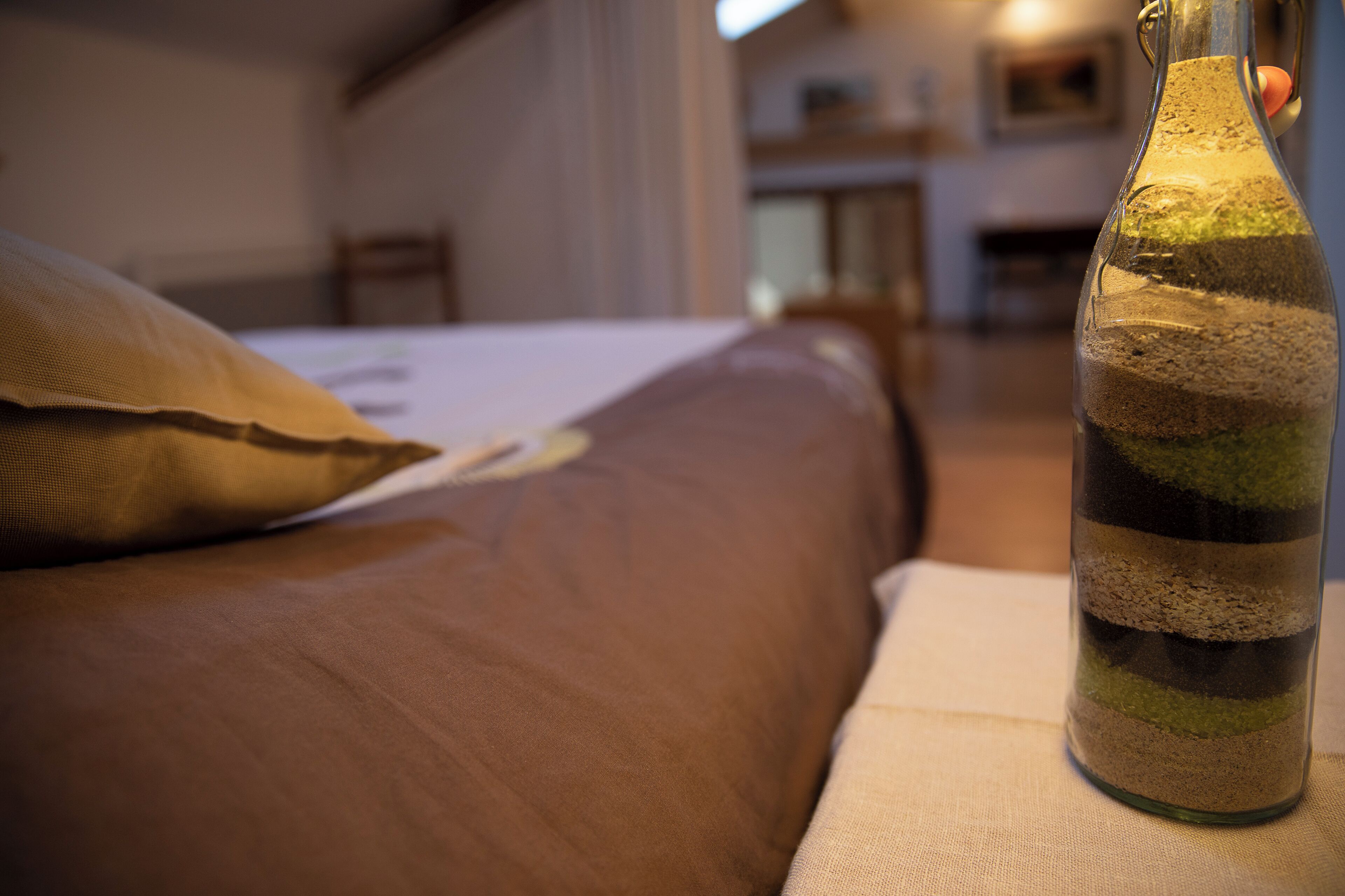 A bed with a brown cover and a pillow, a glass bottle with layered contents, and a wooden chair in a room with a neutral color scheme.