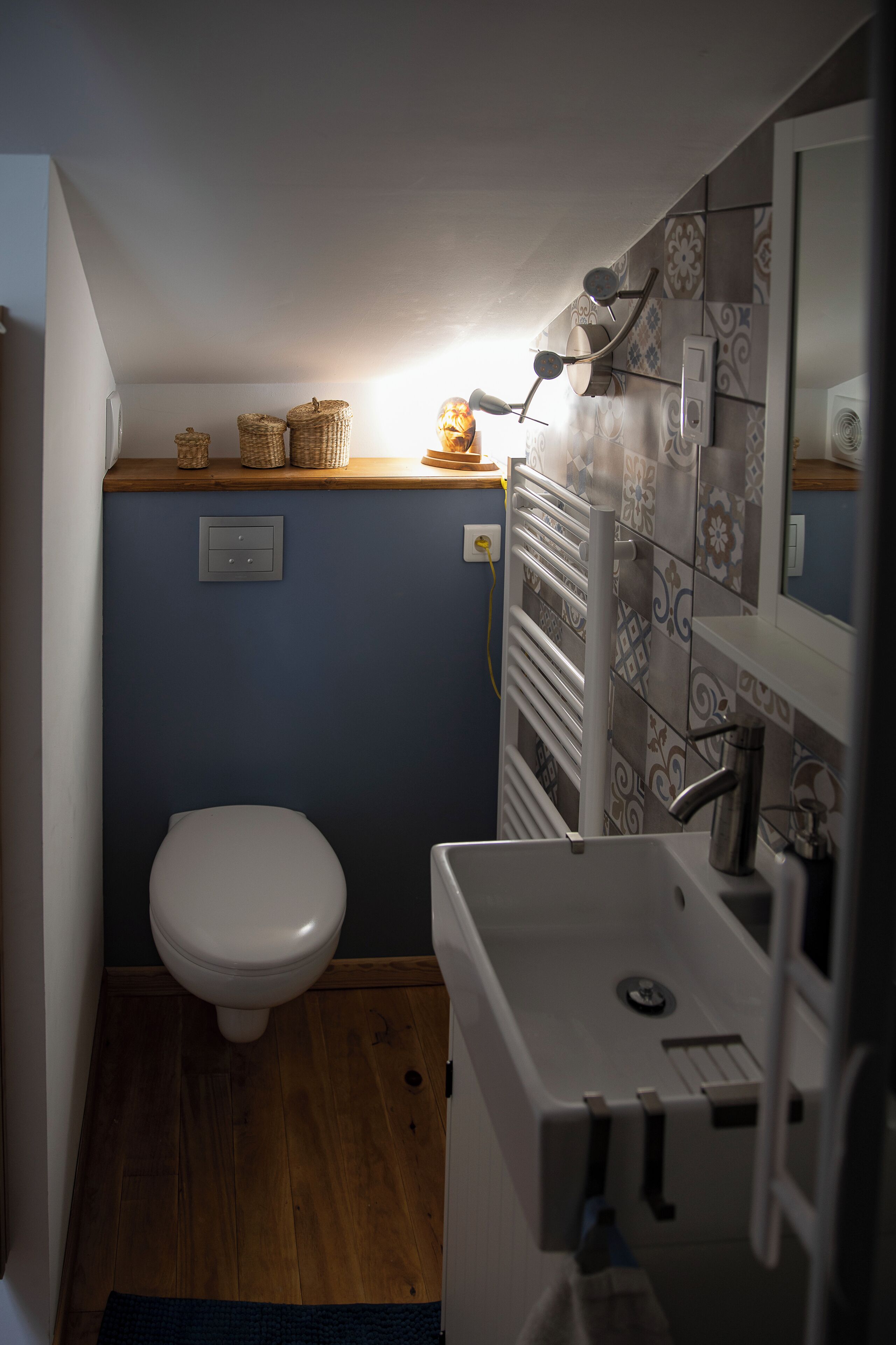 Une petite salle de bain avec des toilettes blanches, un lavabo et un miroir. On y trouve des paniers en osier accrochés au mur et un porte-serviettes.