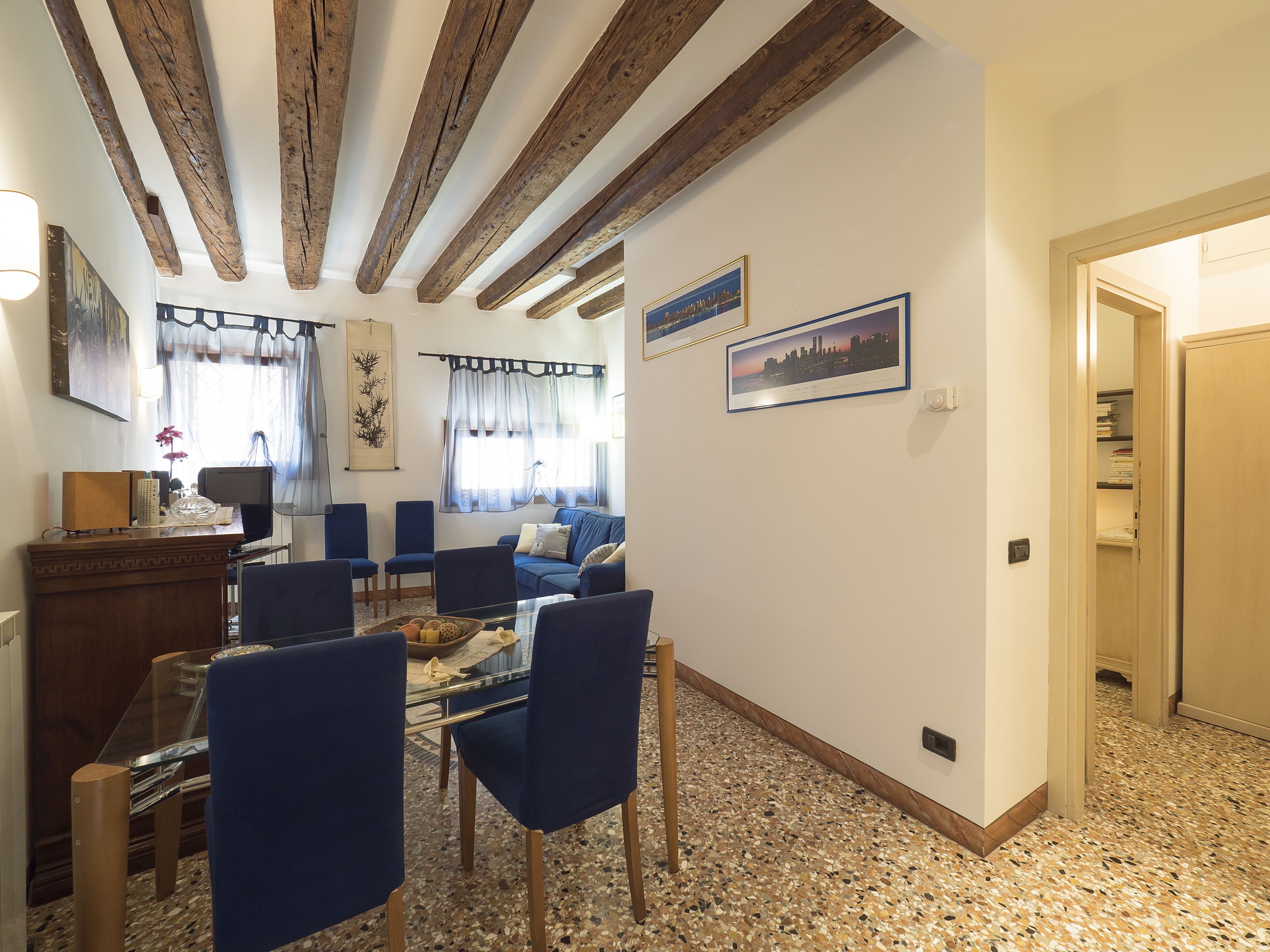 Un salon avec une table en verre, des chaises bleues, un canapé, des poutres en bois au plafond et des tableaux encadrés au mur.