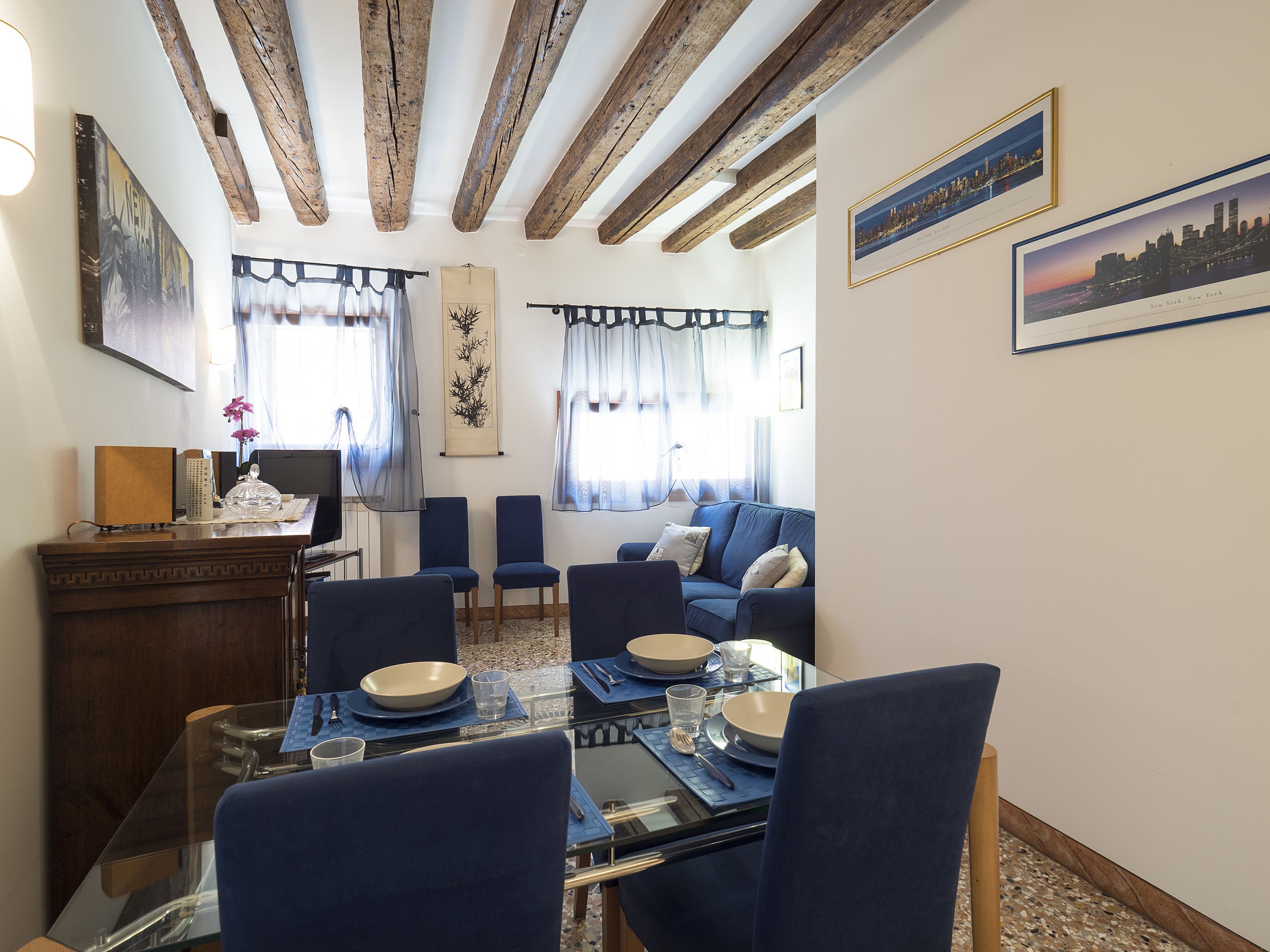 Un comedor con mesa de vidrio, sillas azules y techo de madera.