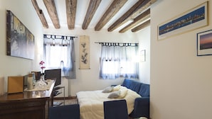 Un dormitorio con cama, un escritorio y una silla. Hay cortinas en las ventanas y un marco de fotos en la pared.