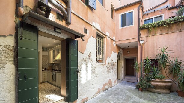 Un patio estrecho con una cocina visible a través de una puerta abierta, suelo de piedra y plantas en macetas.