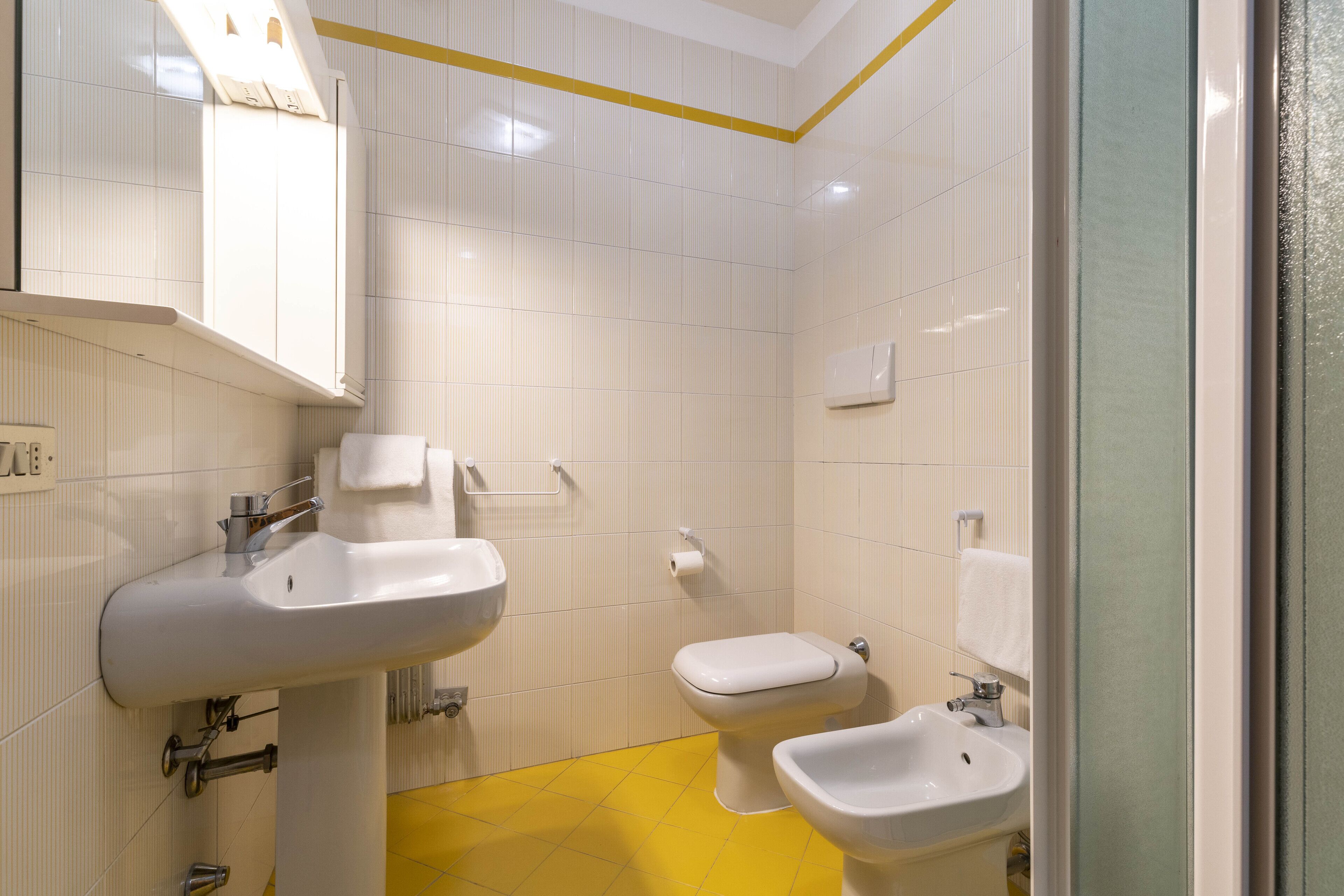 A bathroom with a white sink, toilet, and bidet, yellow floor tiles, and a mirror with a yellow border.