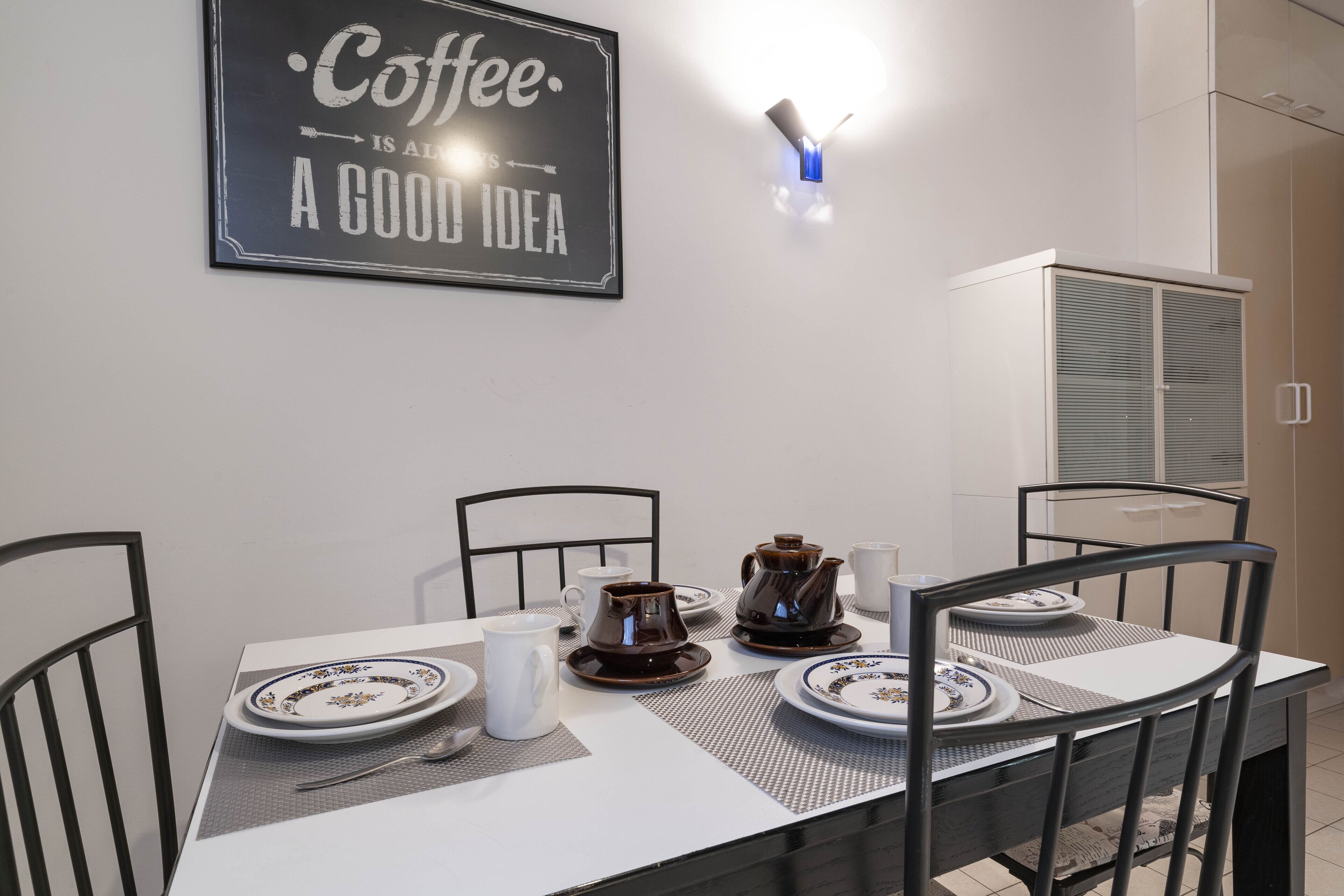 A dining area with a table set for two, featuring plates, cups, and a coffee pot. A sign on the wall reads "Coffee is always a good idea."