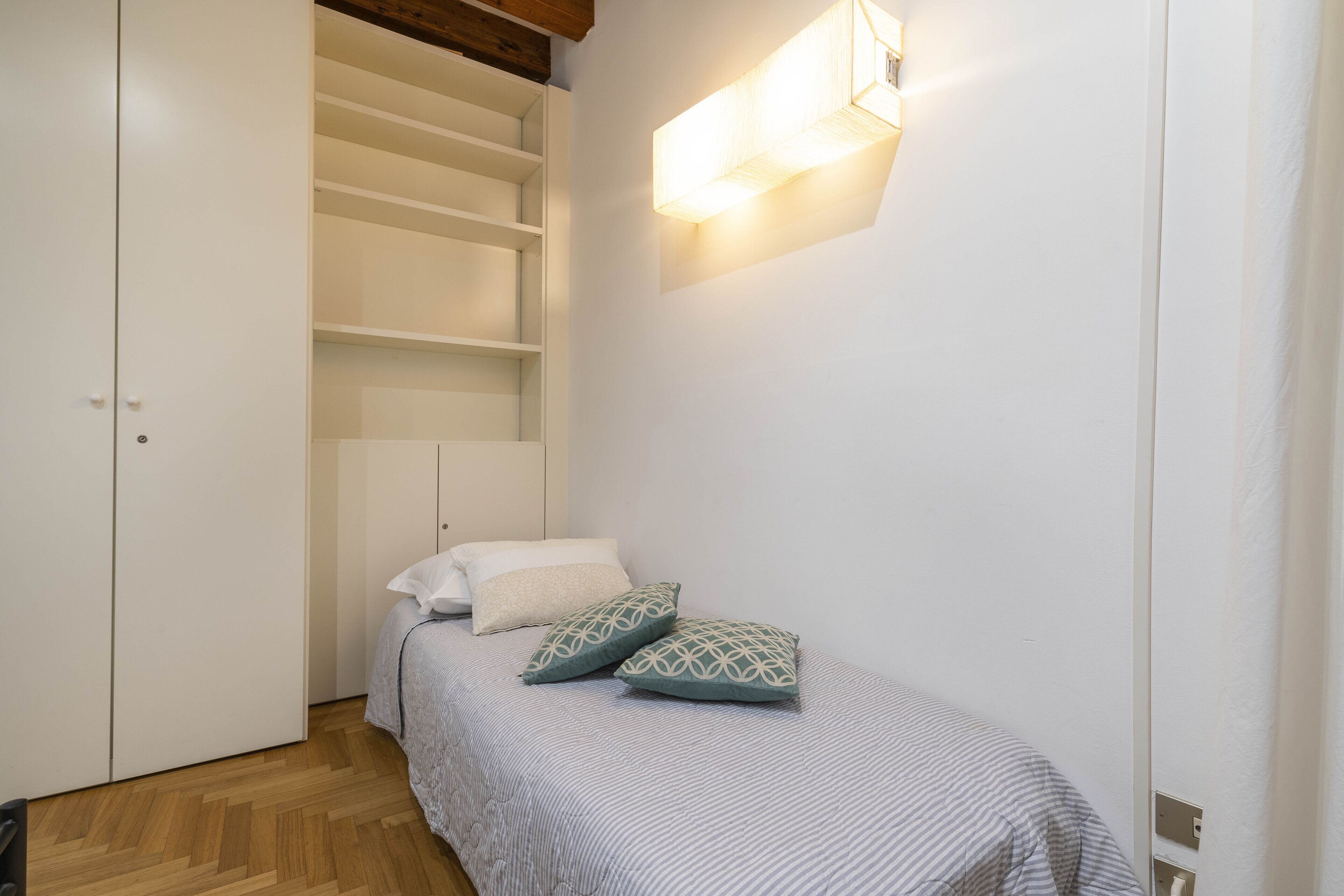 A small, neatly organized bedroom with a bed, two pillows, a nightstand, and a built-in wardrobe.