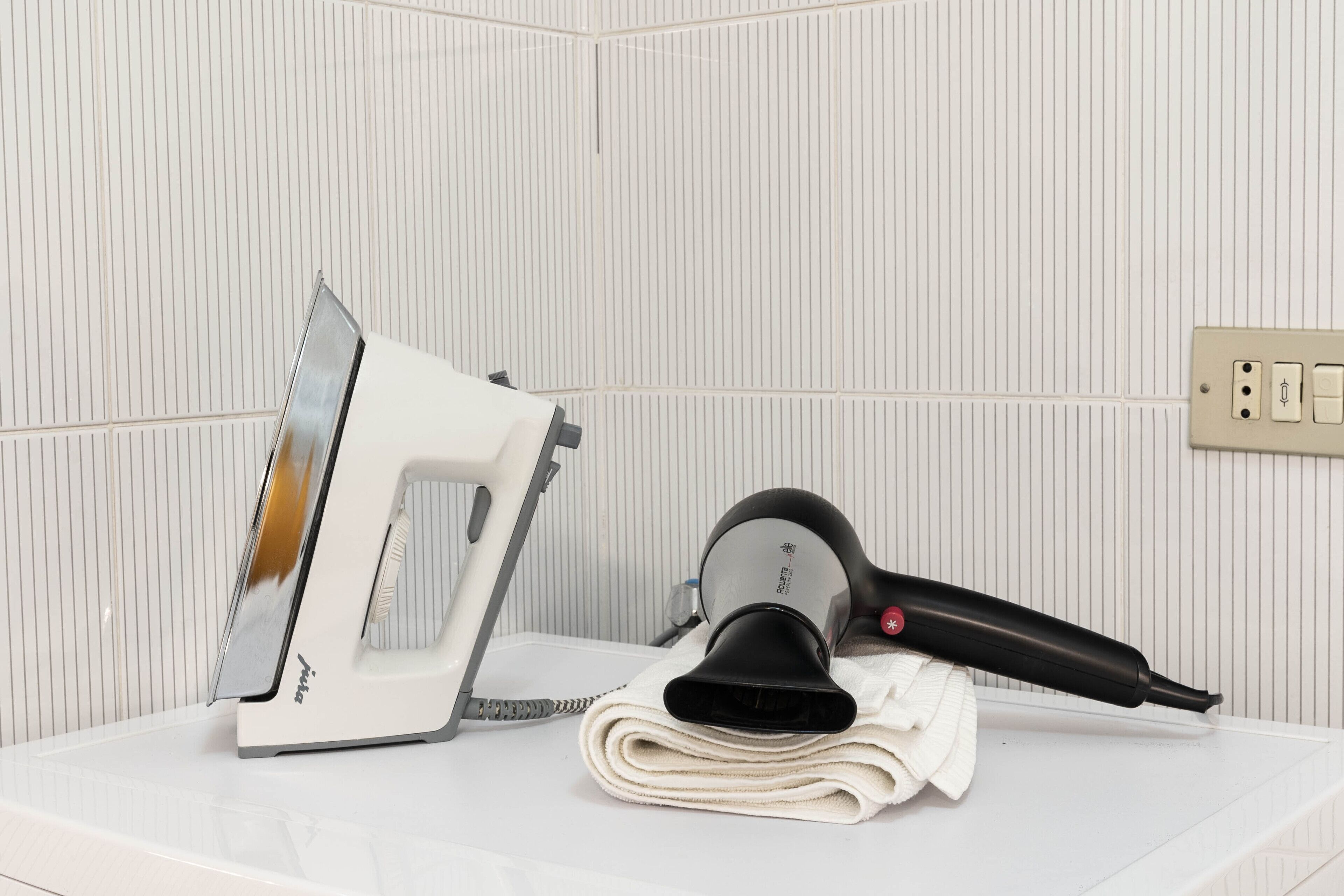 A bathroom with a white countertop, a white iron, a black hairdryer, and a towel.