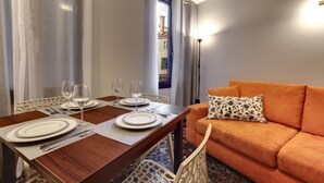 A dining area with a wooden table set for two, a sofa with a floral pillow, and a view of a building through a window with curtains. - San Marco Superior Apartments by Wonderful Italy (Venice)