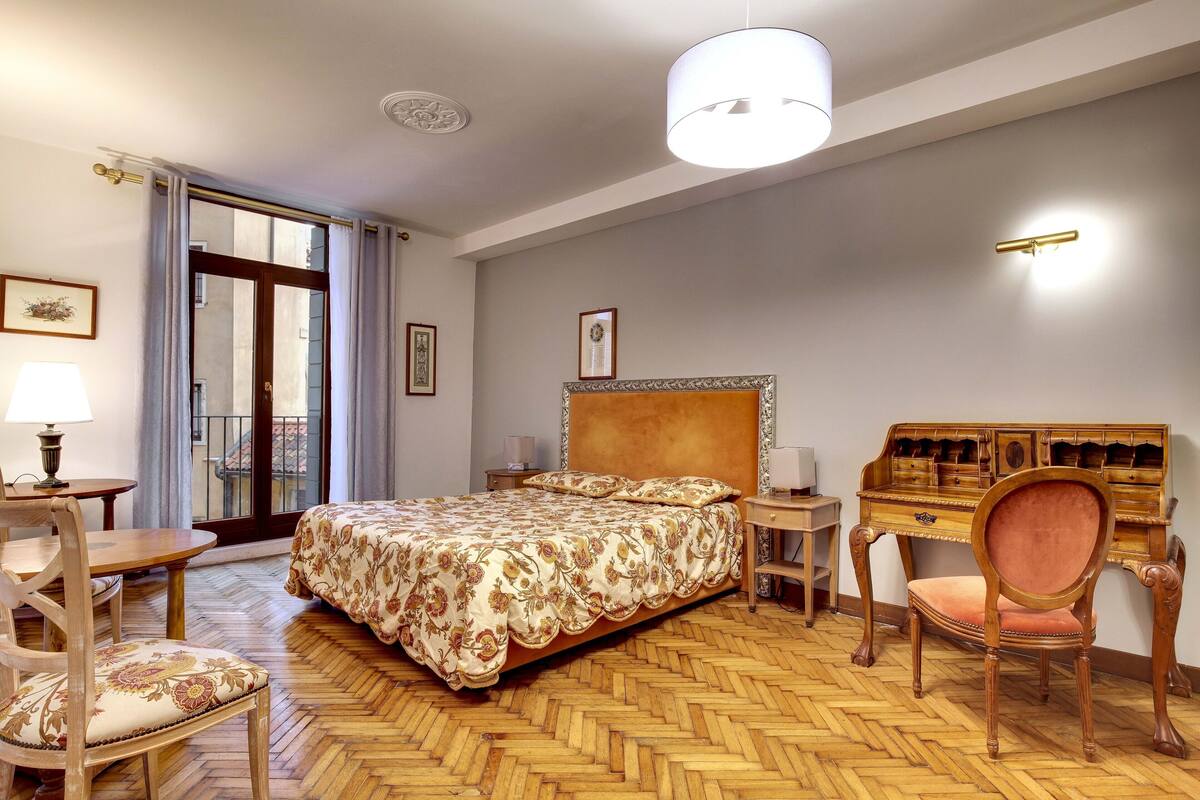 Ein Schlafzimmer mit Holzboden im Fischgrätmuster, einem großen Bett mit blumigem Überwurf, einem Schreibtisch mit Schubladen, einem Stuhl und einer Lampe.
