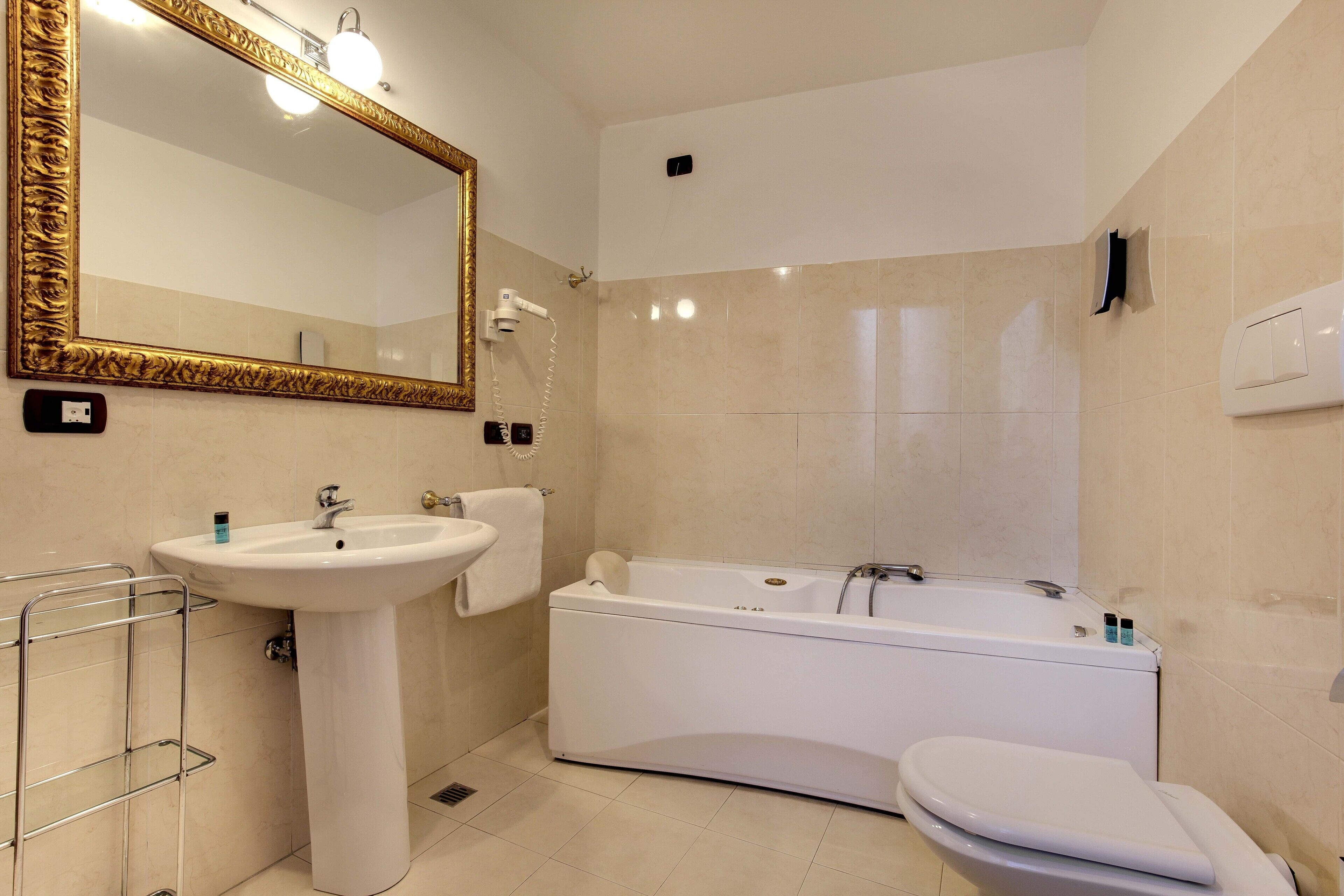 A bathroom with a pedestal sink, a large mirror with a gold frame, a bathtub, and a toilet.