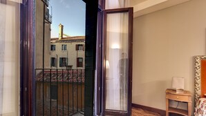 A room with a wooden herringbone floor, a small wooden nightstand with a lamp, and a view of a building with a tiled roof through an open door. - San Marco Superior Apartments by Wonderful Italy (Venice)