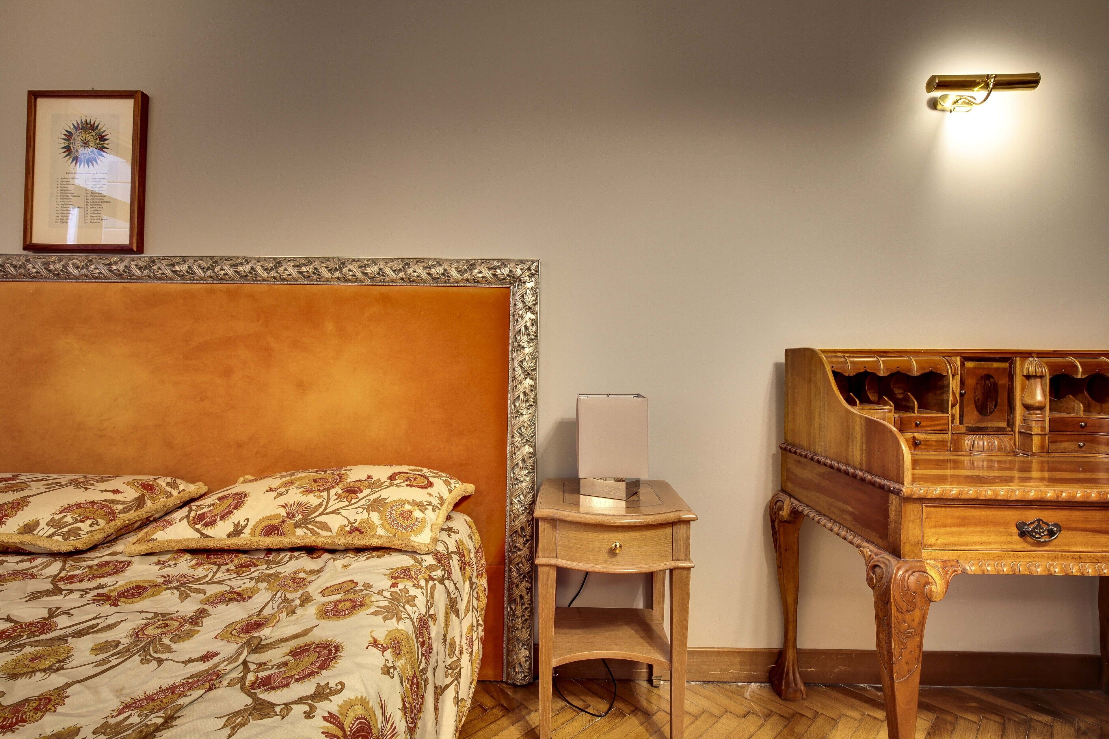 A bedroom with a bed, a wooden nightstand, a desk with drawers, a framed picture on the wall, and a wall-mounted light fixture.