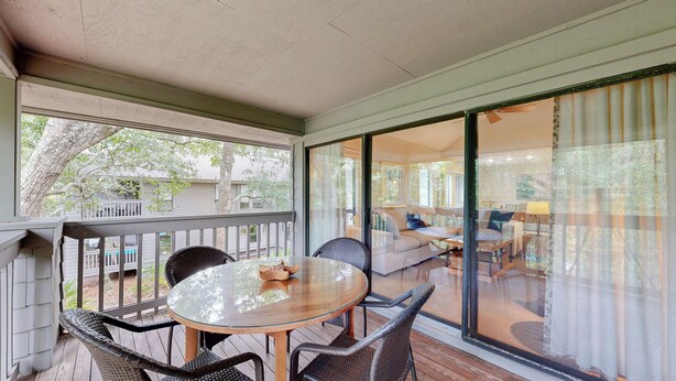 Un espacio exterior techado con una mesa de vidrio y sillas de mimbre, con vistas a una sala de estar con sofá y lámpara.