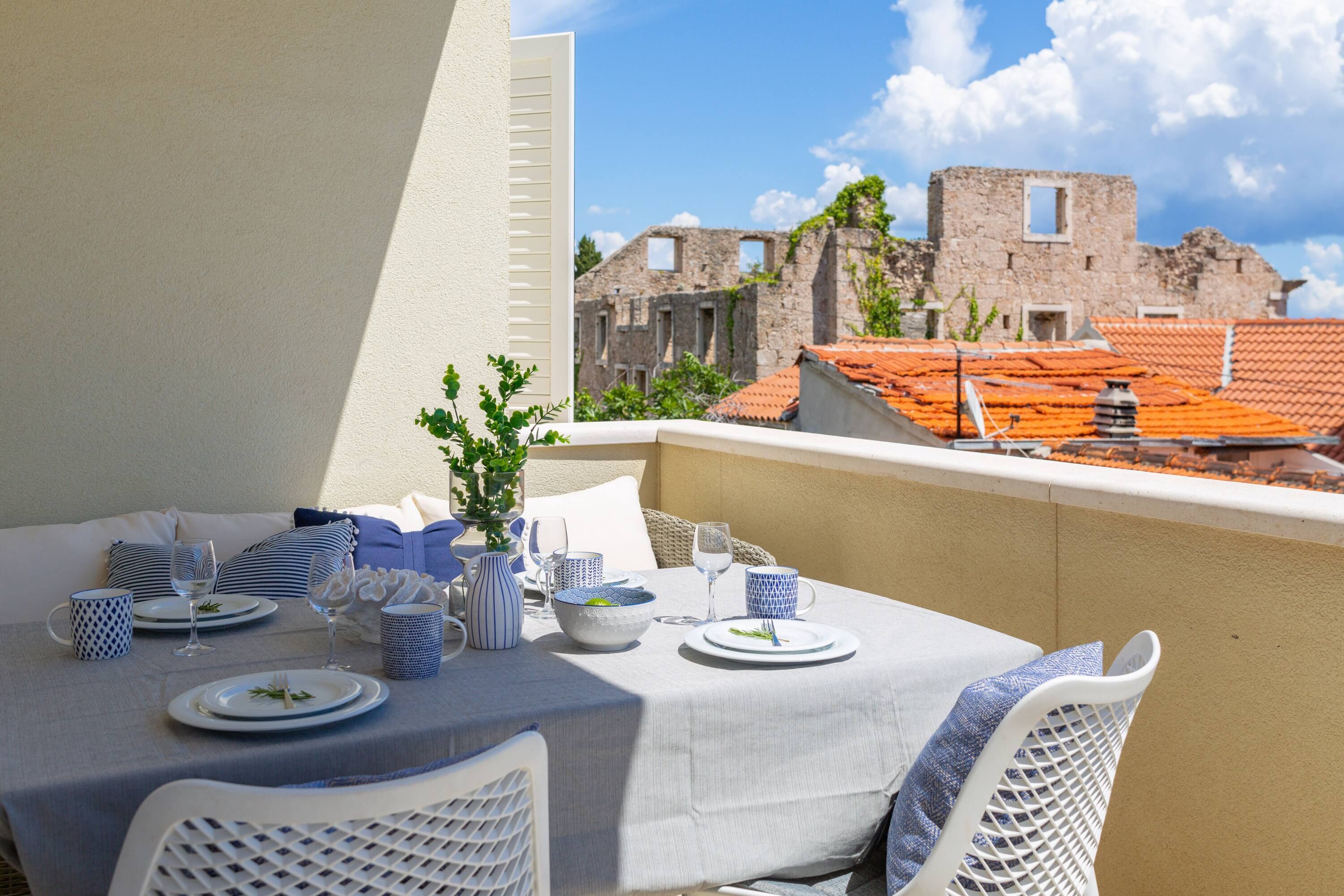 Un balcon avec une table dressée pour le dîner, des chaises en osier blanc et une vue sur des bâtiments historiques en pierre, coiffés de toits en tuiles rouges.