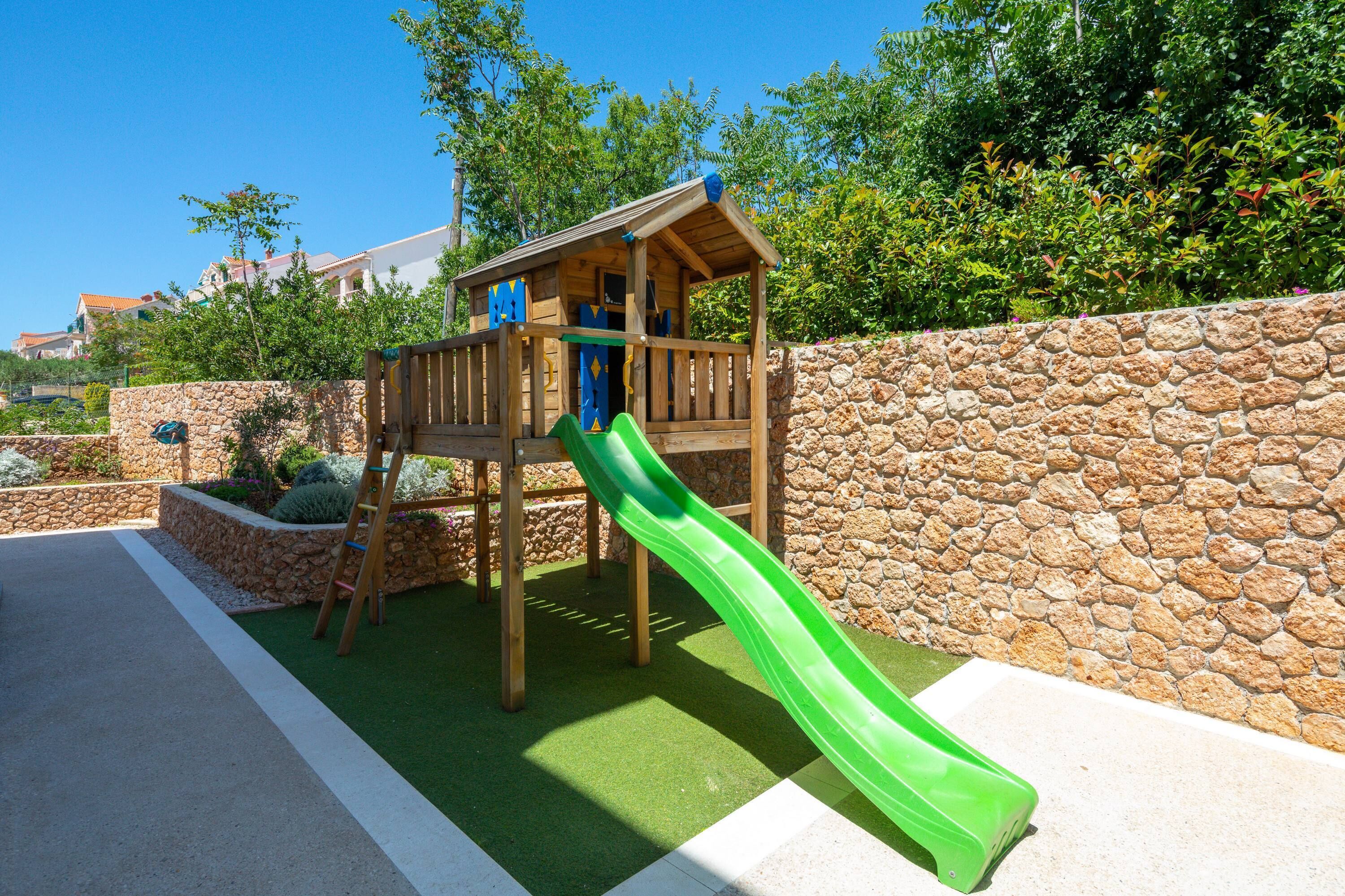 Una struttura giochi in legno con uno scivolo verde e una scala da arrampicata, circondata da un muro in pietra e vegetazione.