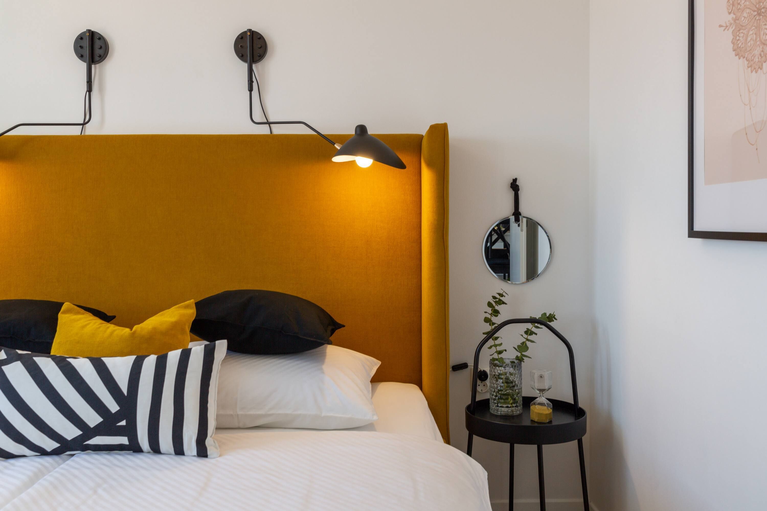 Un lit bien fait, agrémenté d’un coussin rayé, d’un coussin jaune et d’un coussin noir. On y trouve une table de chevet avec un miroir, une petite plante et une bougie.