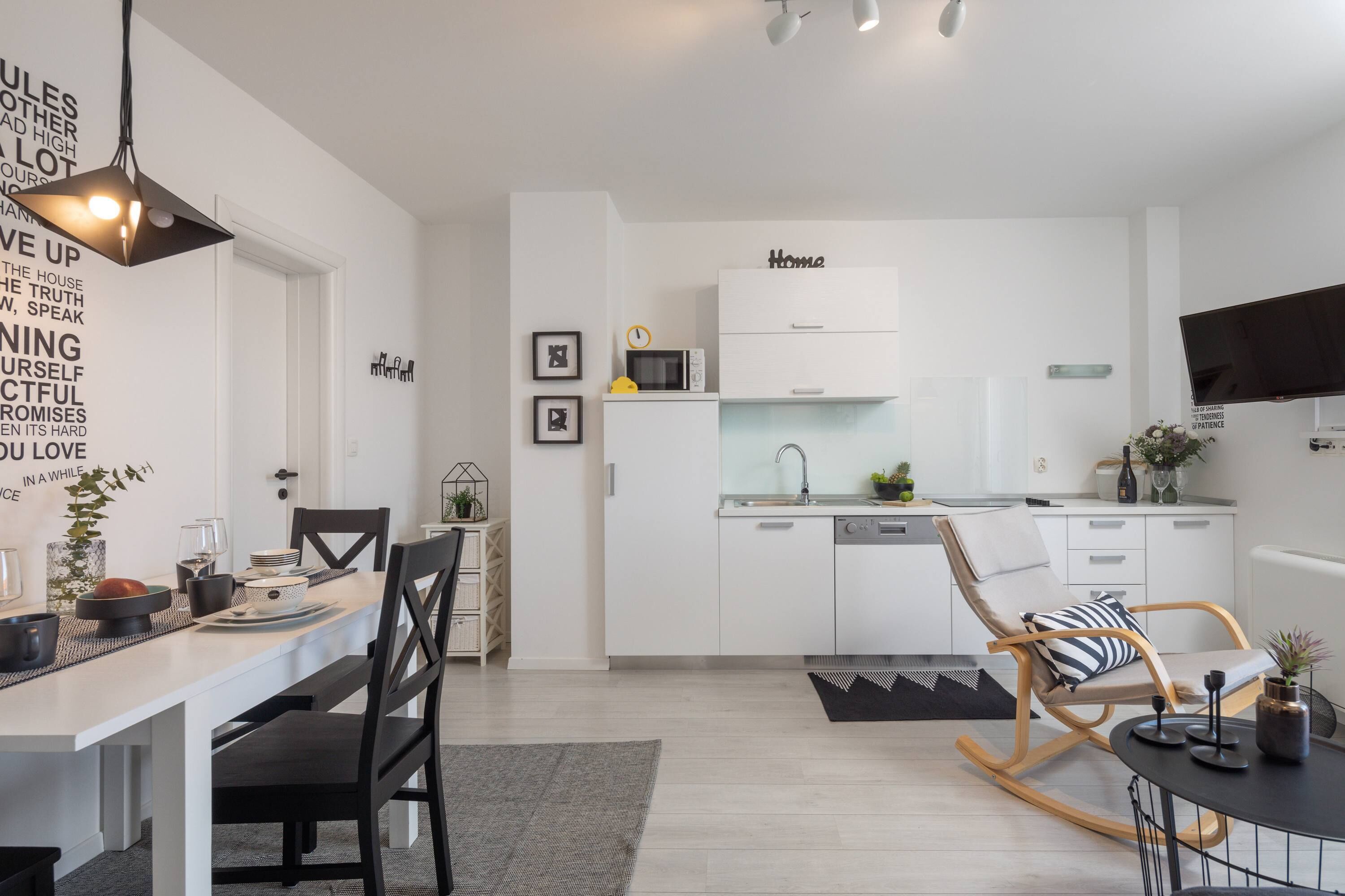 A modern, minimalist kitchen with white cabinetry, a dining area with a white table and black chairs, and a living area with a black sofa and a wooden rocking chair.