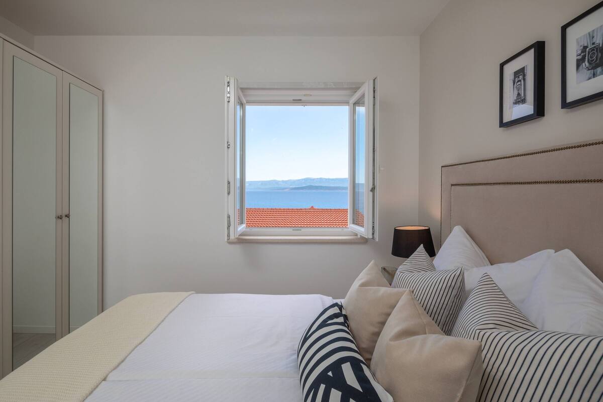 A bedroom with a bed, pillows, a nightstand, a lamp, a window with a view of the sea, and a wardrobe.