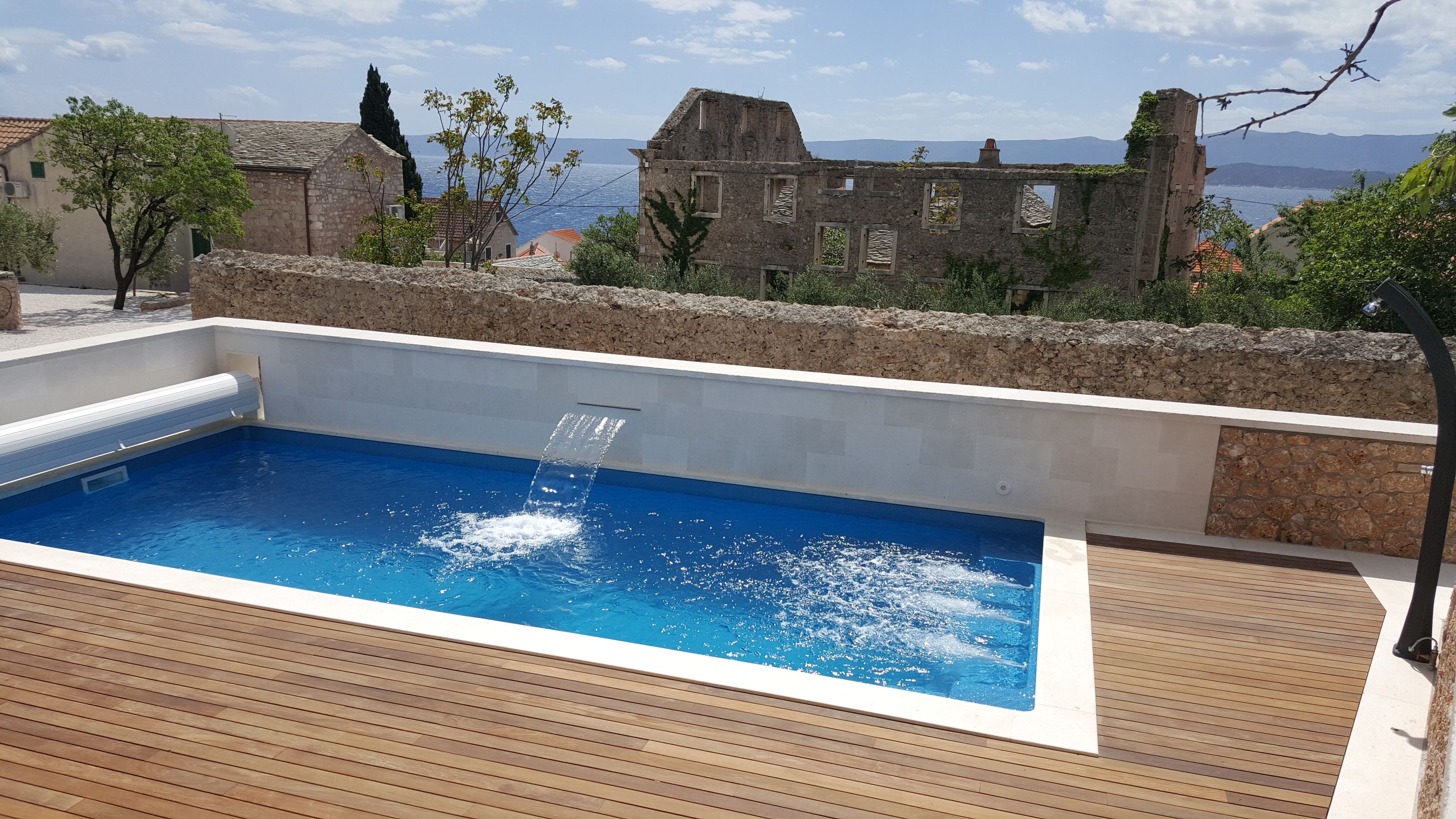 A swimming pool with a waterfall feature, surrounded by stone walls and wooden decking, overlooking a coastal landscape with historic buildings.