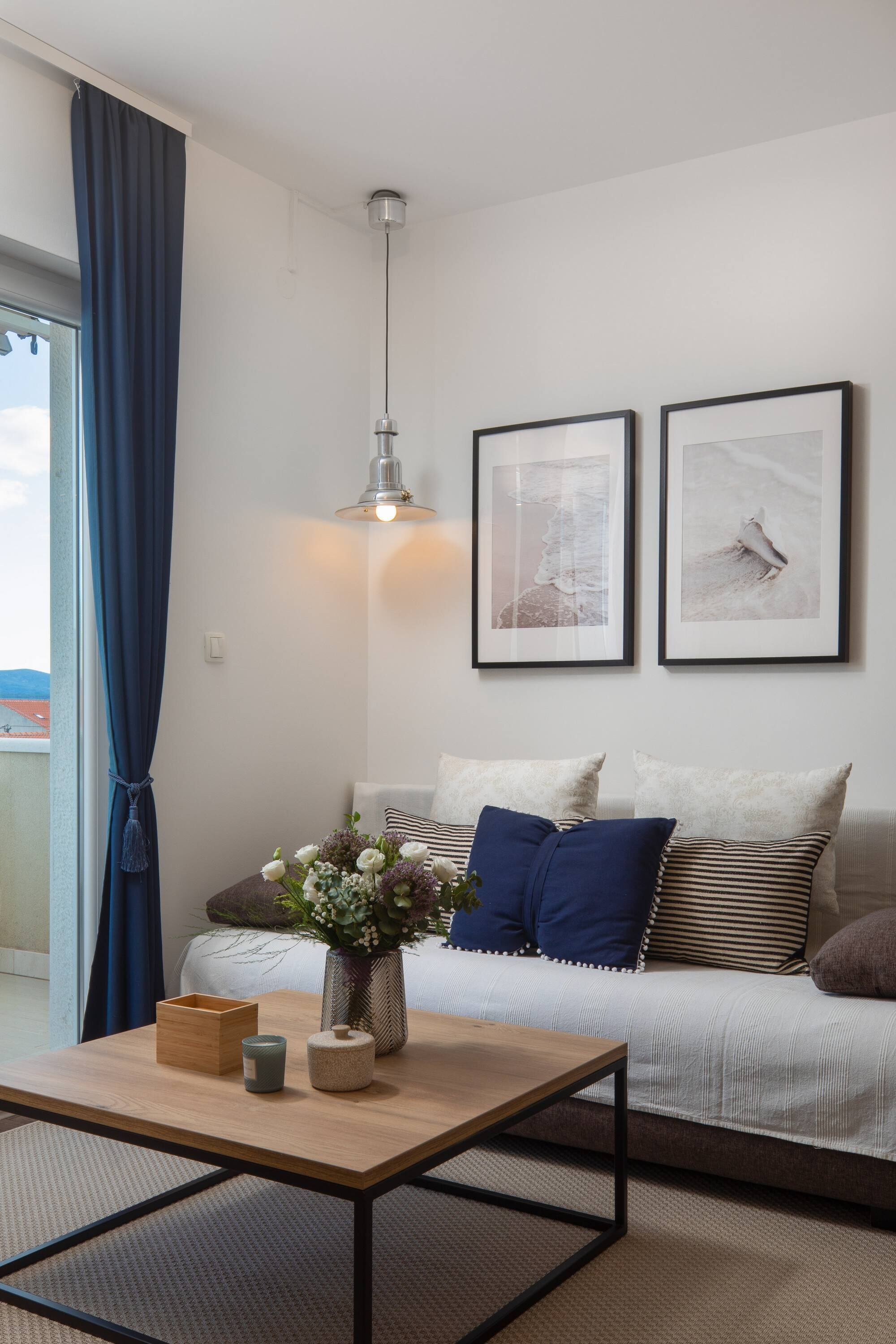 A modern living room with a sofa, a coffee table, and a vase with flowers. There are two framed pictures on the wall and a window with curtains.