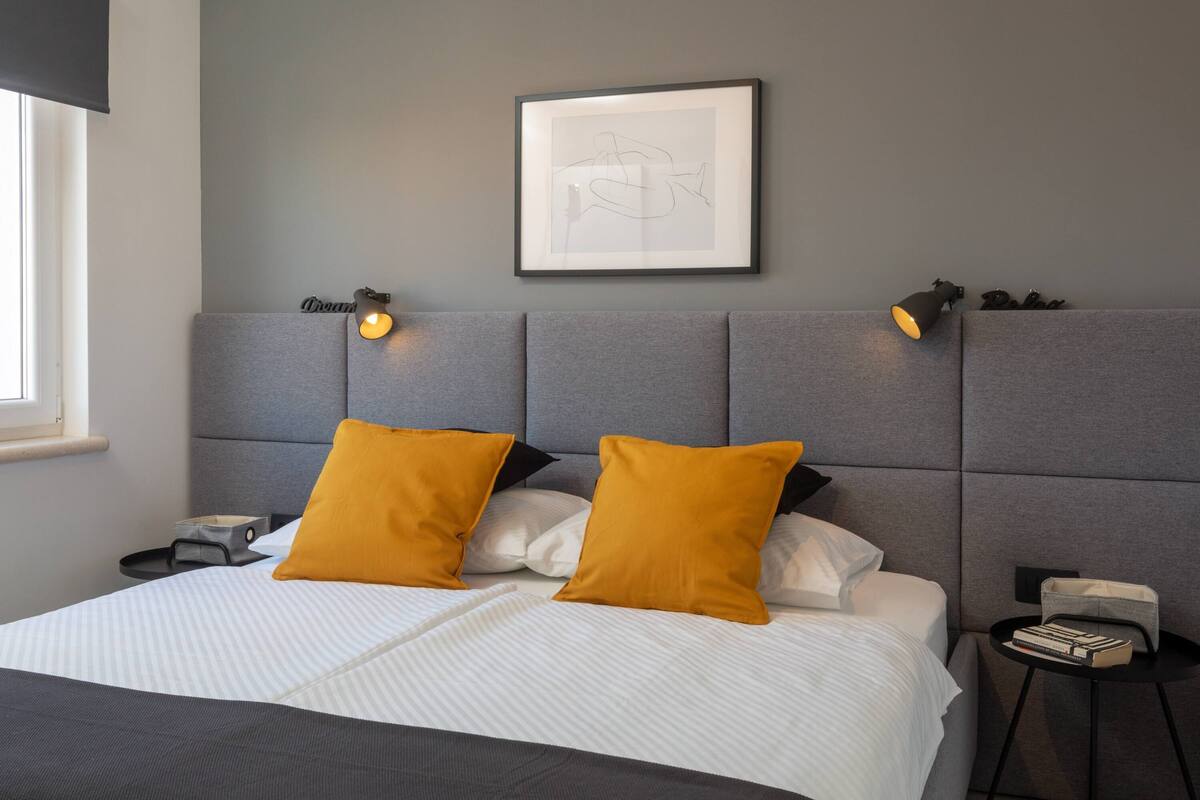 A modern bedroom with a grey upholstered headboard, a framed artwork above the bed, and a bedside table with a lamp and books.
