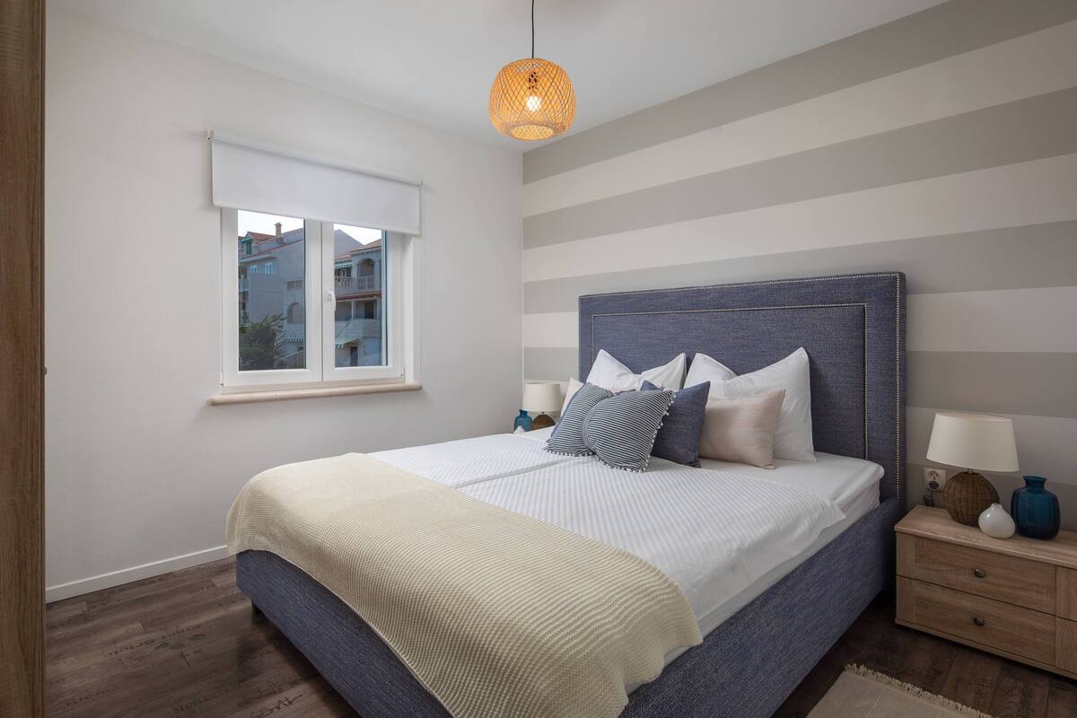 A bedroom with a large bed, a window with blinds, a bedside table with a lamp, and a hanging light fixture.