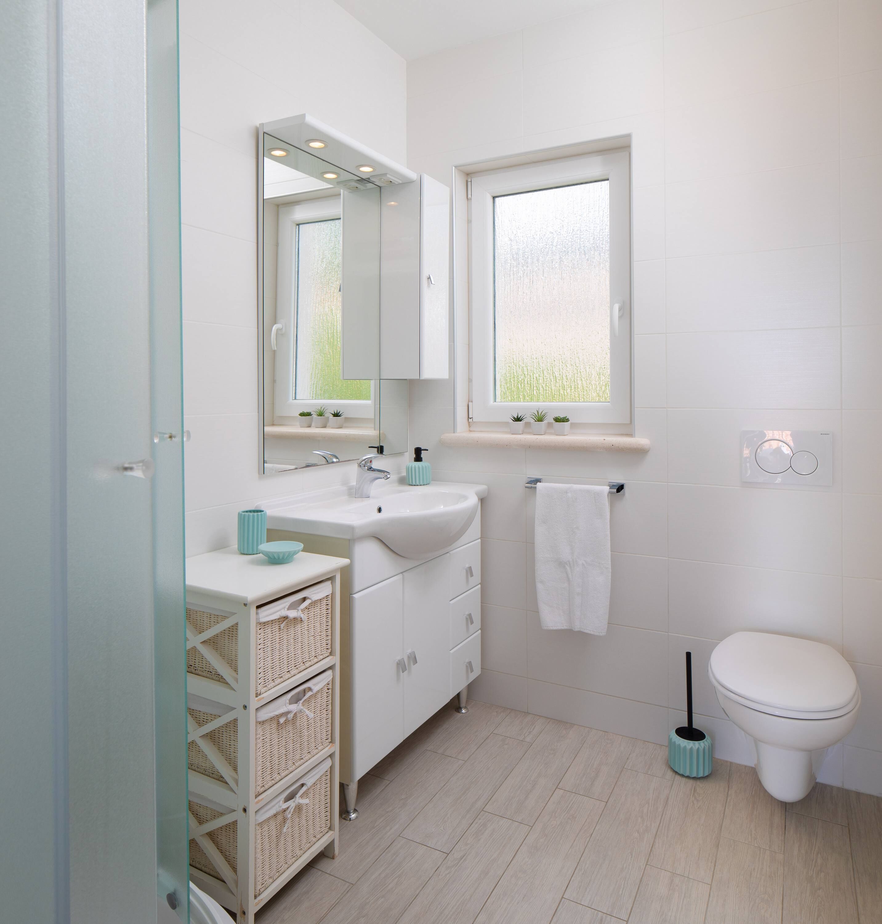 Un baño moderno con lavamanos, espejo y inodoro blancos. Hay cestas de mimbre debajo del lavamanos y una ventana sobre el inodoro.