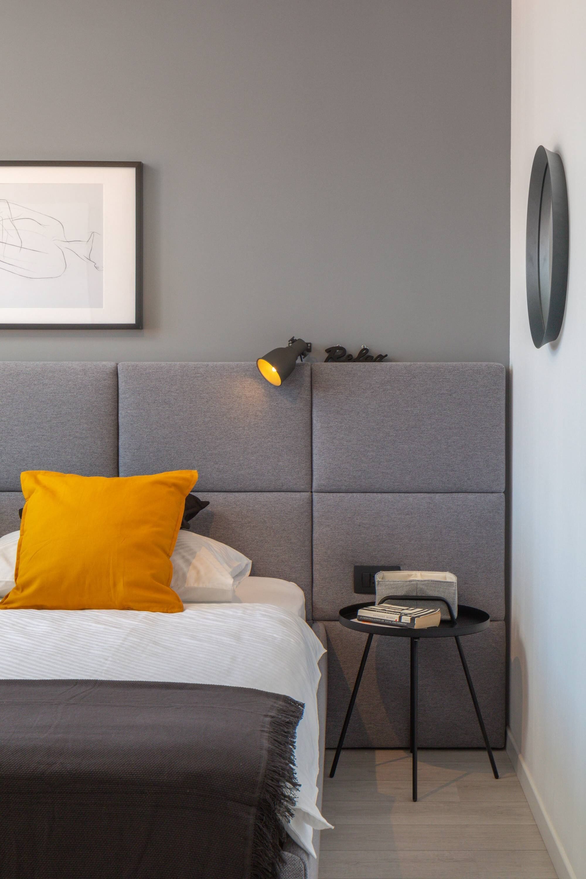 A modern bedroom with a grey headboard, a bedside table with books, and a framed artwork on the wall.