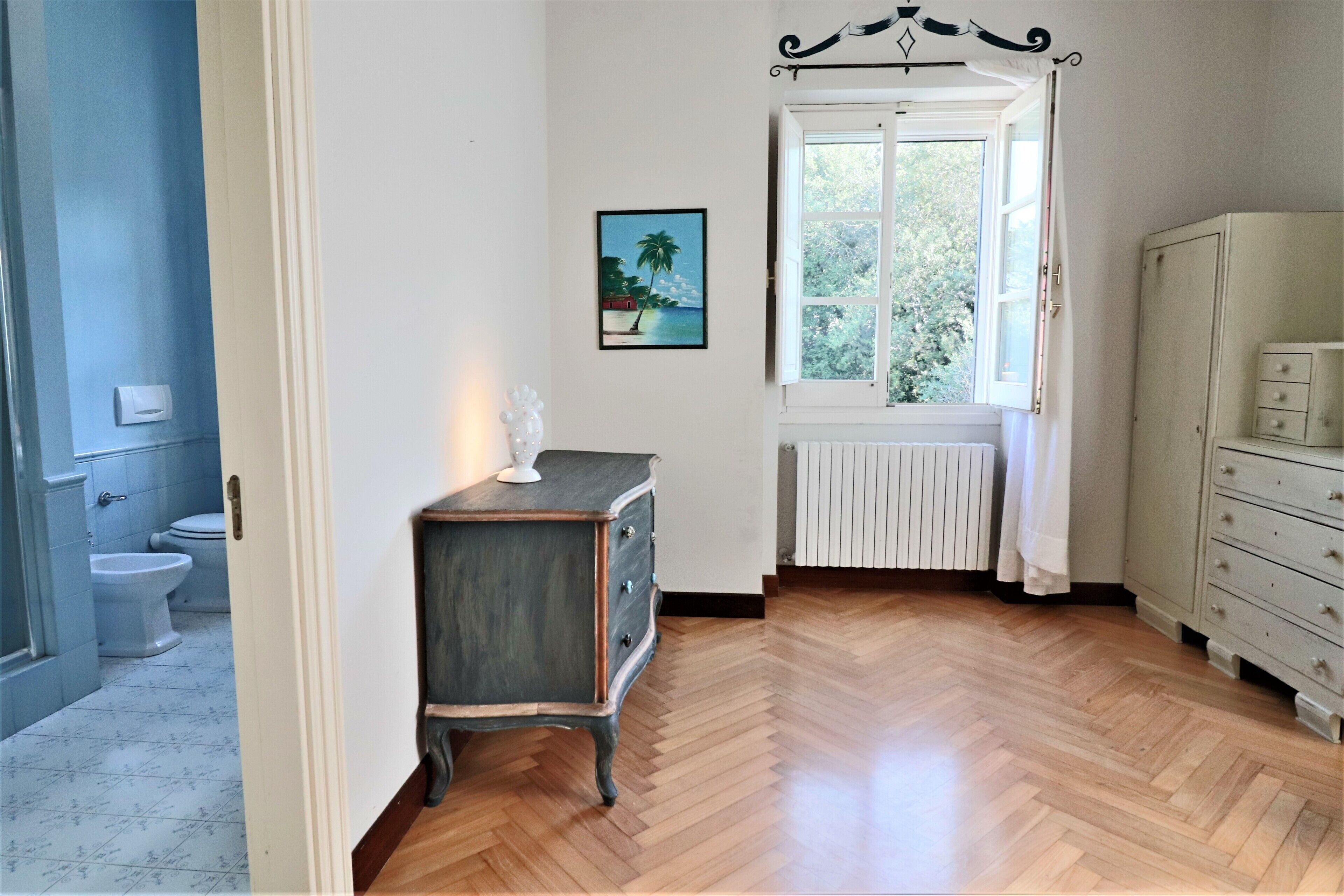 Una habitación con suelo de madera en patrón espiga, un radiador blanco, una ventana con cortinas blancas, un aparador blanco y un cuadro que muestra una escena de playa.