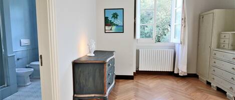 Una habitación con suelo de madera en patrón espiga, un radiador blanco, una ventana con cortinas blancas, un aparador blanco y un cuadro que muestra una escena de playa.