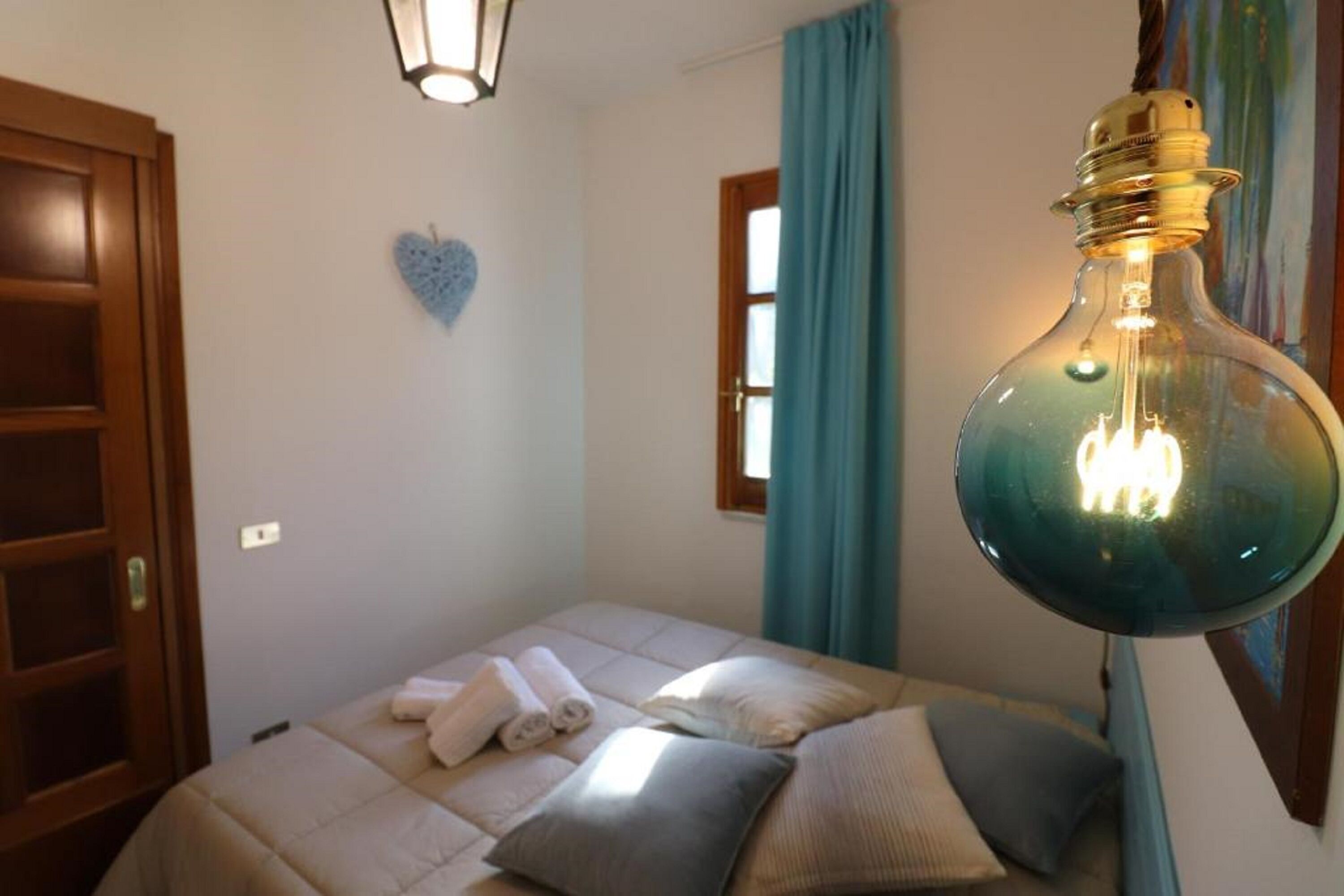 Camera da letto con un letto, asciugamani, una decorazione a forma di cuore, una finestra con tende e una lampada a sospensione.