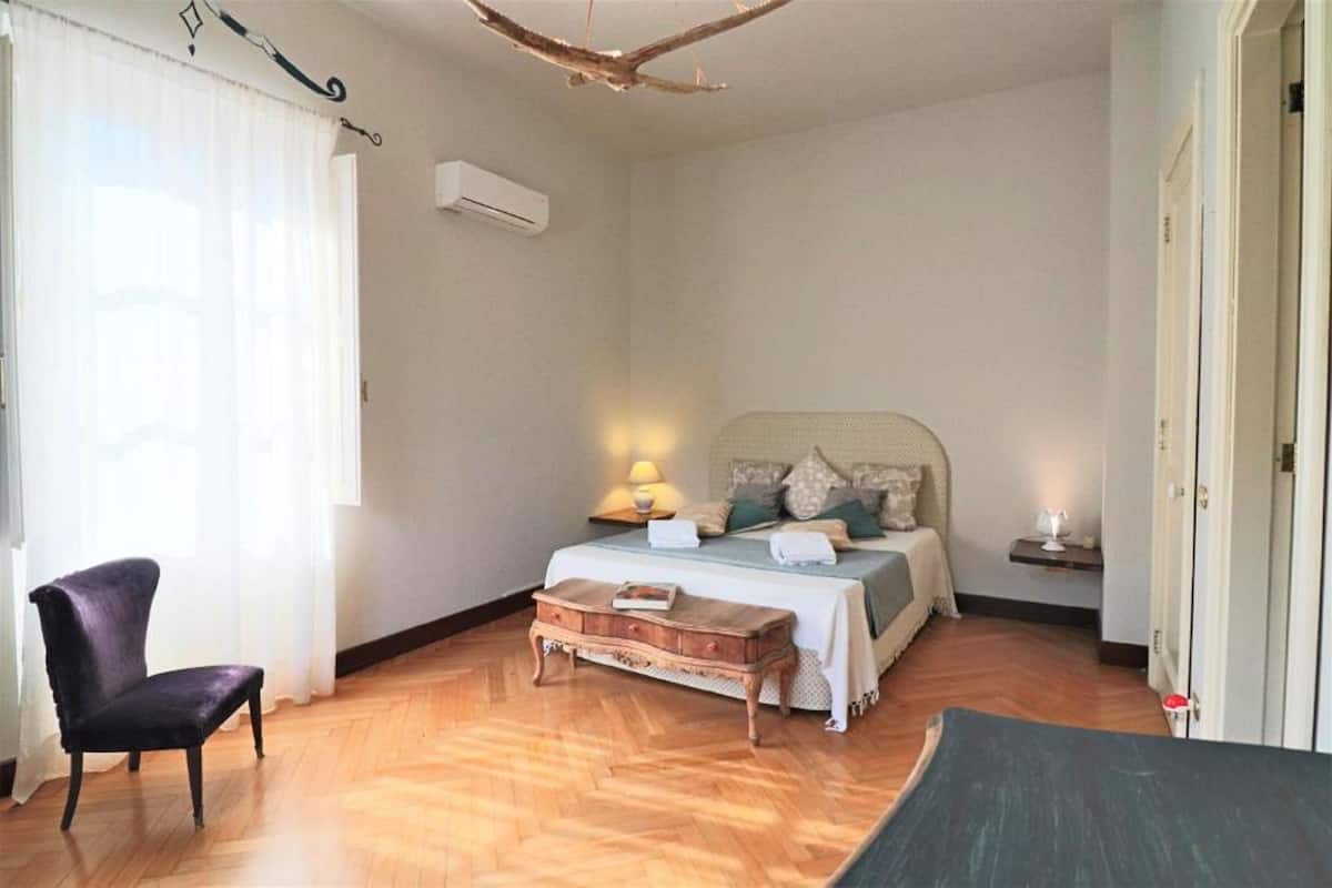 A bedroom with a bed, a chair, a wooden bench, and a bedside table with a lamp.