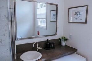 A bathroom with a white sink, a large mirror, a framed drawing on the wall, and a potted plant.