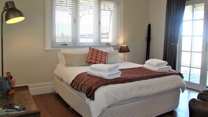 Ein ordentlich gemachtes Bett mit weißer Bettwäsche, einem rot-weißen Kissen und einer Streifen-Decke. Eine Lampe befindet sich auf der linken Seite des Bettes, im Hintergrund ein Fenster mit Jalousien.