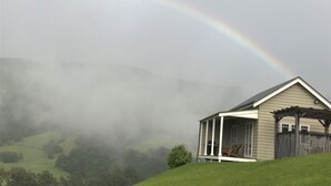 Ein Haus mit einer Veranda und einem Holzzaun, umgeben von einem grasbewachsenen Hügel und einem Regenbogen am Himmel.