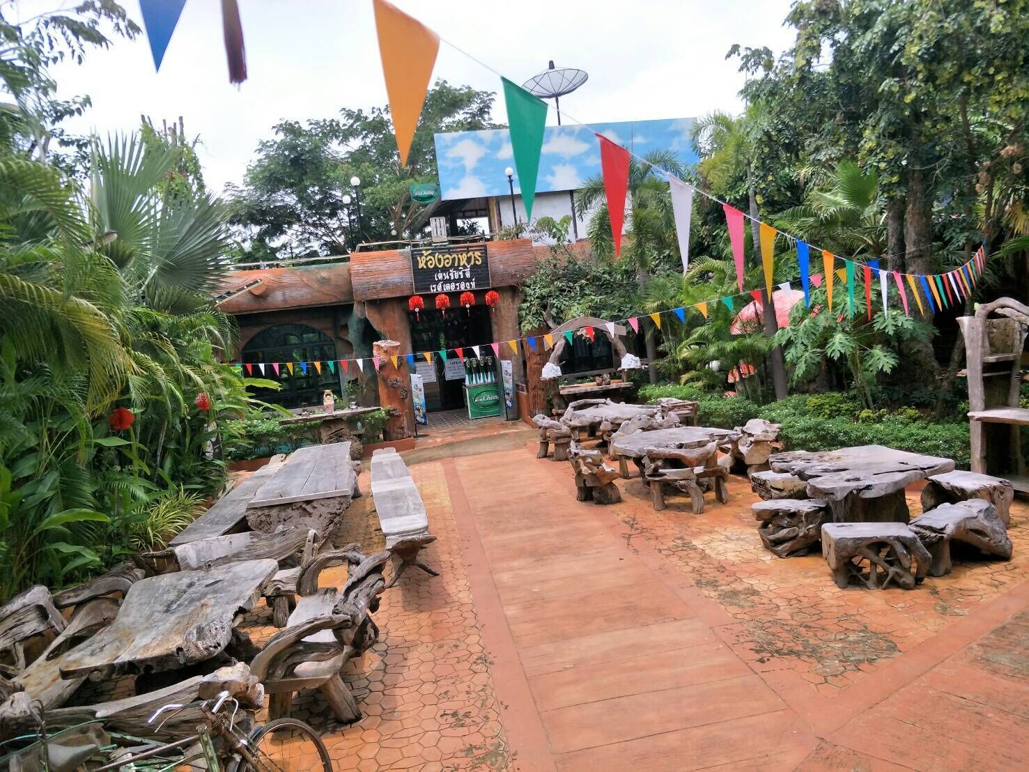 An outdoor seating area with rustic wooden tables and benches, surrounded by lush greenery and a building with a sign in a foreign language.