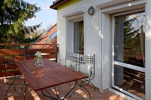 Eine Außenpatio mit einem Holztisch und Metallstühlen, eine weiße Wand mit einem Fenster und einem Metallgeländer.