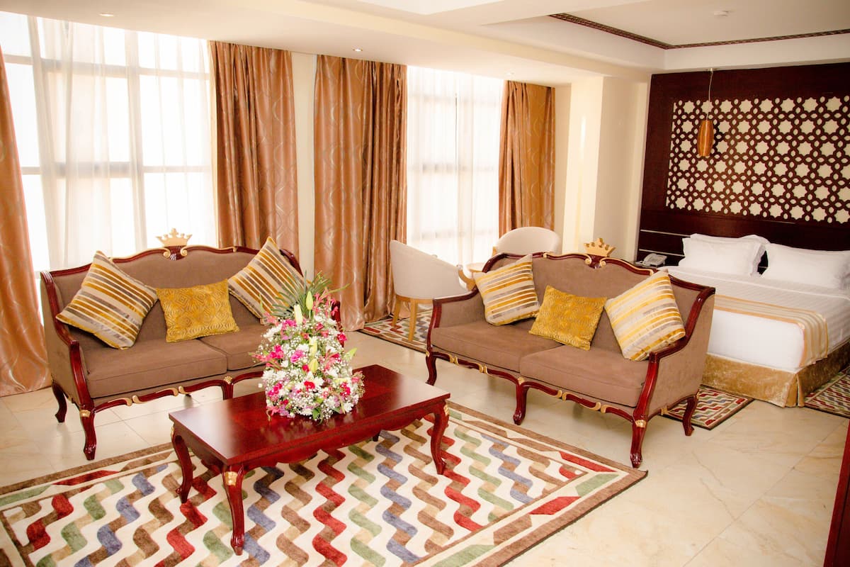 A hotel room with a king-sized bed, two sofas with decorative pillows, a wooden coffee table with a floral arrangement, and a patterned rug.