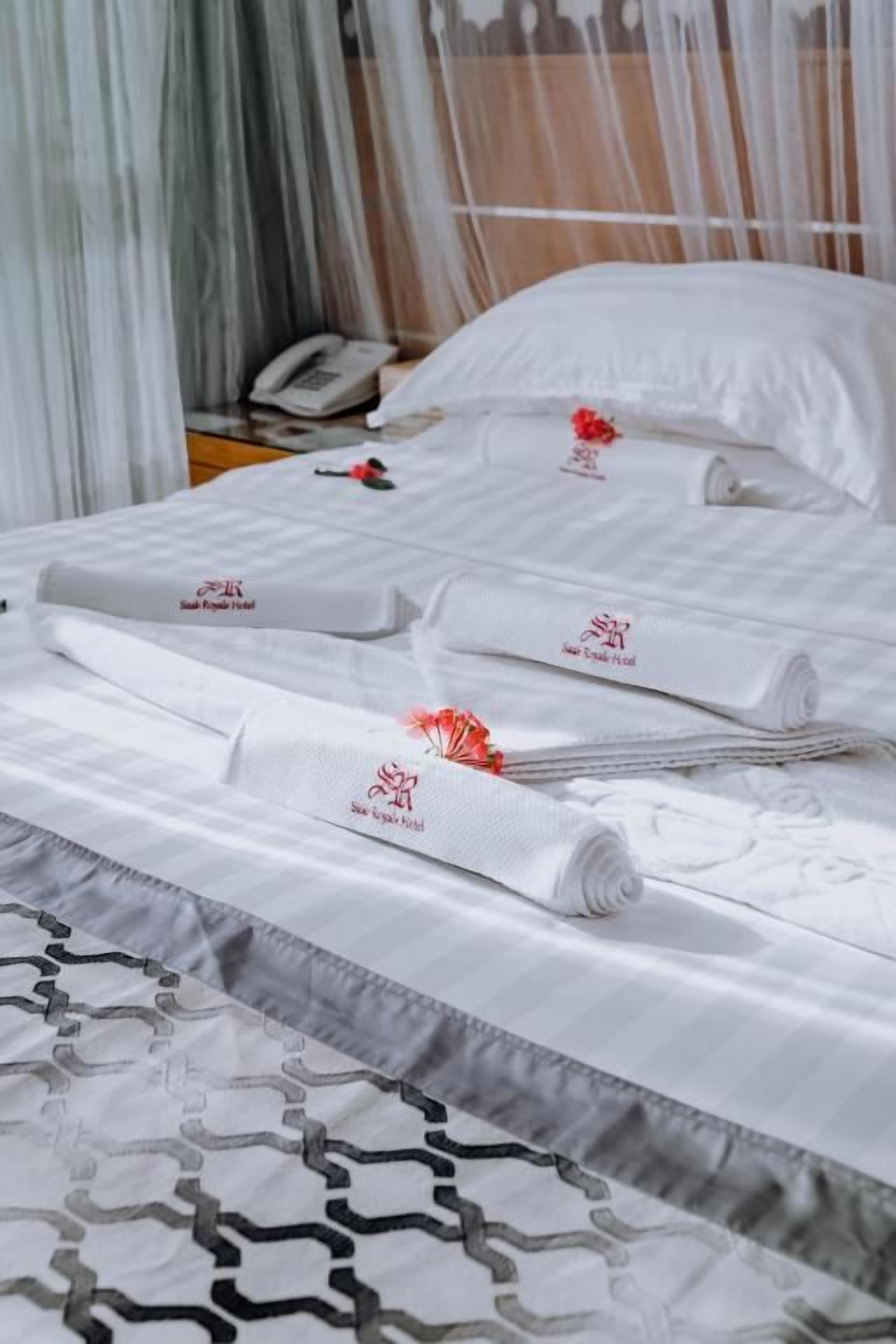 Una cama bien hecha con toallas blancas enrolladas y una decoración de flor roja, un teléfono blanco en la mesita de noche y una colcha estampada.