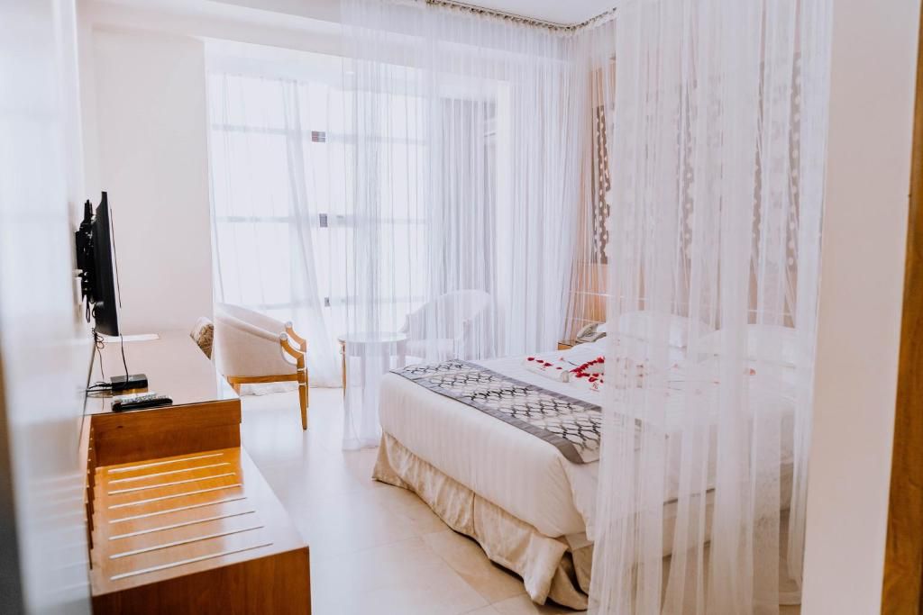 A bedroom with a bed, a desk, a chair, and a window with sheer curtains.