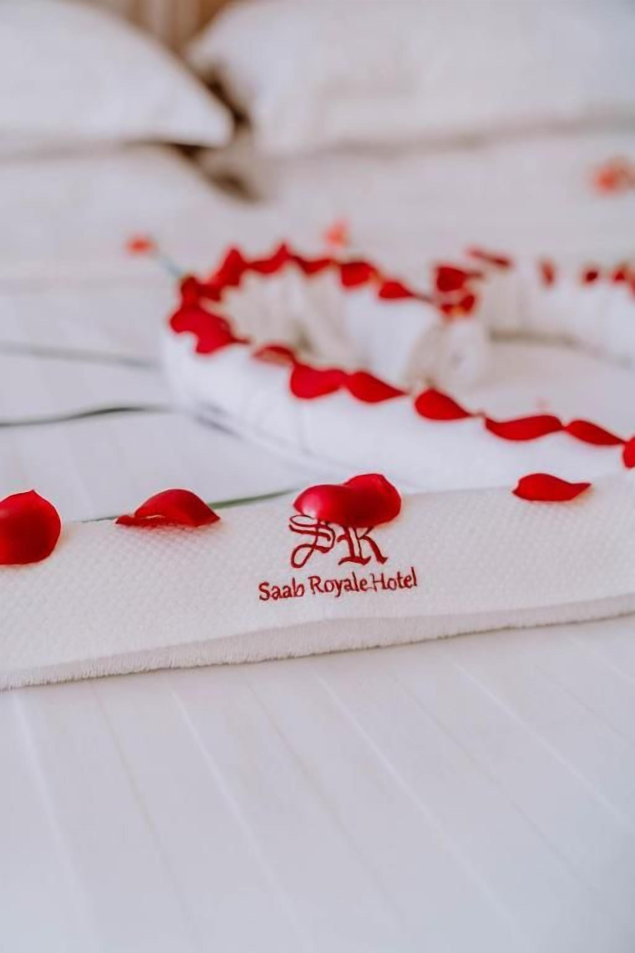 Un lit recouvert d’une courtepointe blanche et orné de pétales de roses rouges disposés en forme de cœur. Le logo « Saab Royale Hotel » est visible sur la courtepointe.