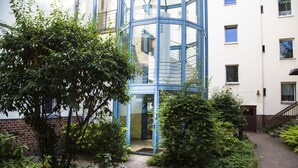 A modern building with a glass elevator shaft, surrounded by greenery and a brick wall. - Hevals Grand Tempelhof (Berlin)
